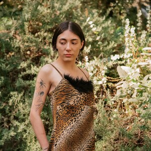 May include: A woman wearing a brown and black animal print mini dress with a black feather trim at the neckline. She is standing in a garden setting.