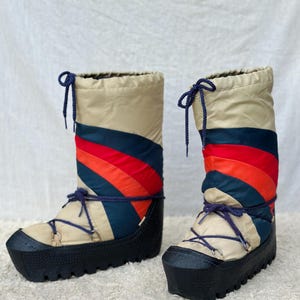 May include: A pair of vintage snow boots. The boots are beige with a colorful striped design in red, orange, and navy blue. They have black soles and navy blue laces. The boots are designed to keep feet warm and dry in snowy conditions.