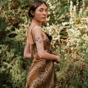 May include: A woman wearing a brown and black animal print slip dress with a black feather trim. She is standing in front of a green leafy background.