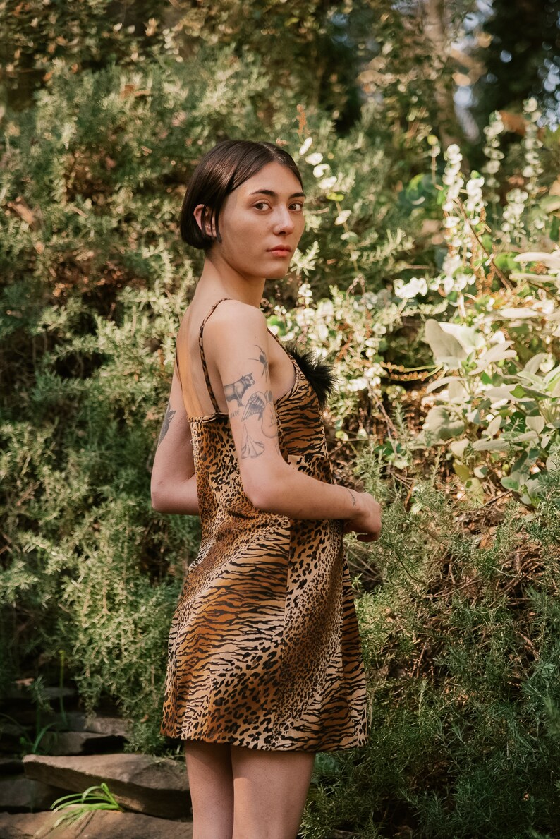May include: A woman wearing a brown and black animal print slip dress with a black feather trim. She is standing in front of a green leafy background.