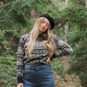 K&ouml;nnte beinhalten: Eine Frau tr&auml;gt eine schwarze Baskenm&uuml;tze, einen schwarz-wei&szlig;en Pullover und eine blaue Jeans. Sie steht in einem Wald.
