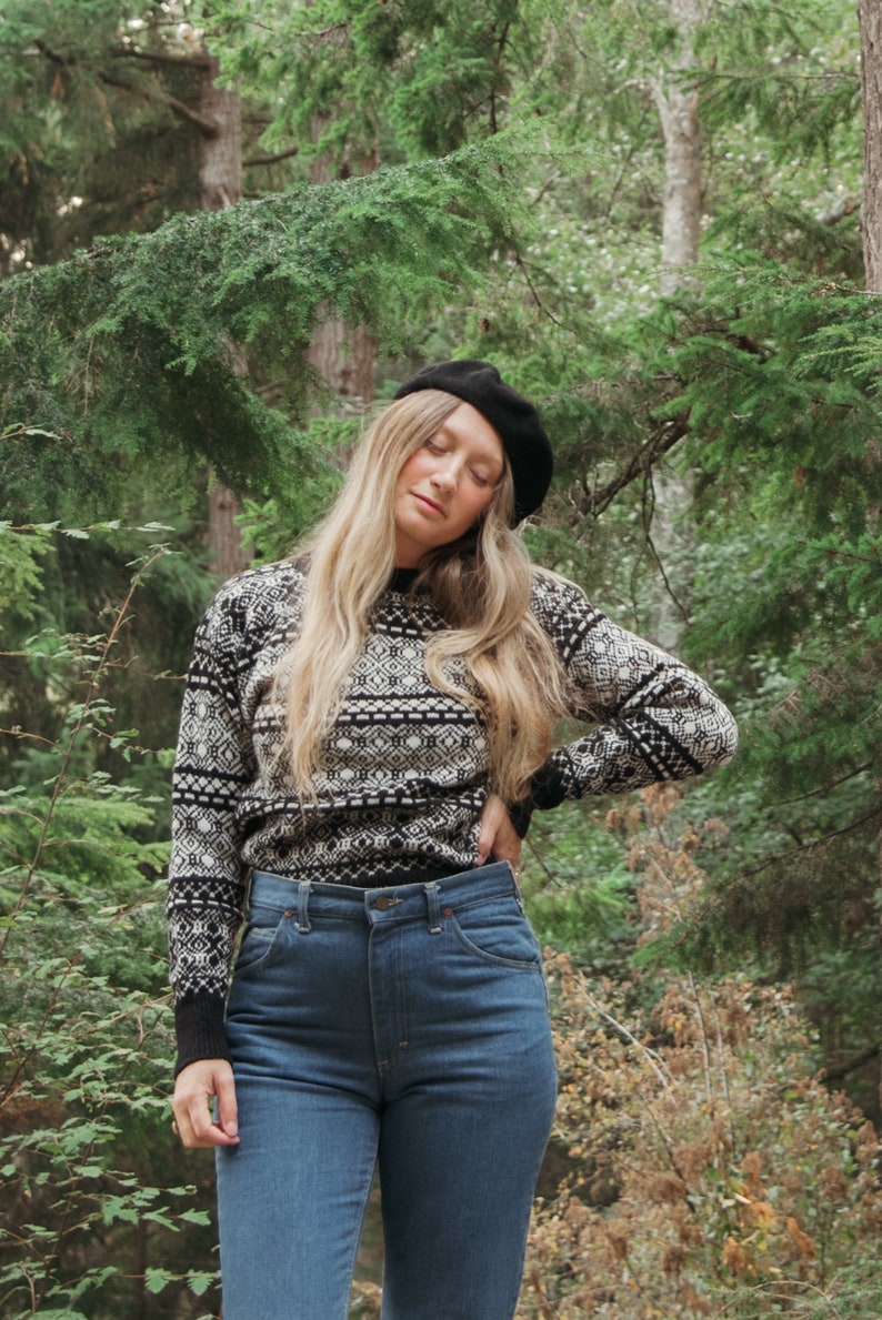 K&ouml;nnte beinhalten: Eine Frau tr&auml;gt eine schwarze Baskenm&uuml;tze, einen schwarz-wei&szlig;en Pullover und eine blaue Jeans. Sie steht in einem Wald.