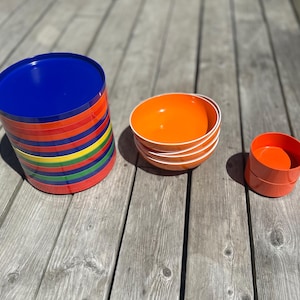 以下が含まれることがあります： 青、赤、オレンジ、黄色、緑のカラフルなプラスチックプレートのスタック。その隣には、白い縁のあるオレンジ色のボウルと、小さなオレンジ色の容器のスタックがあります。アイテムは風化した木製の表面に配置されています。
