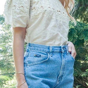 May include: Off-the-shoulder cream-colored top with lace detailing and light blue denim shorts. The shorts have a rolled hem and a Lee label. A gold bracelet is visible on the wrist.
