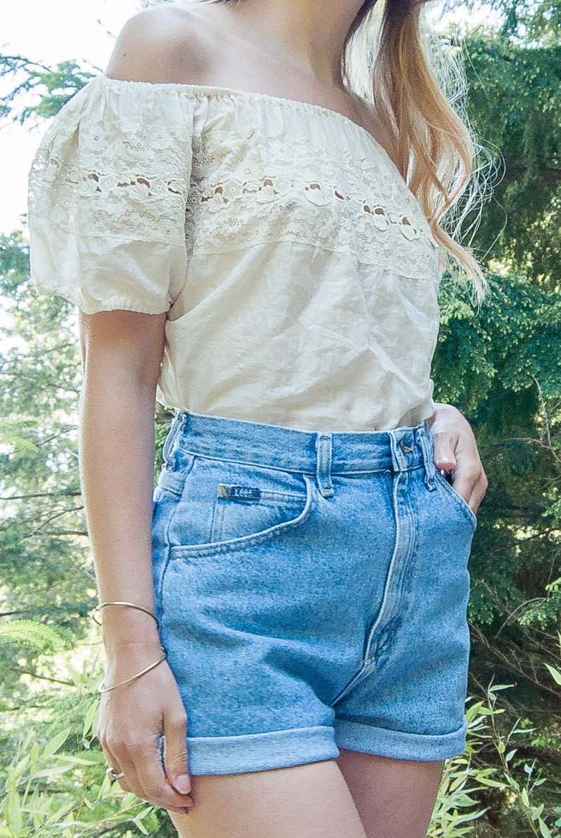 May include: Off-the-shoulder cream-colored top with lace detailing and light blue denim shorts. The shorts have a rolled hem and a Lee label. A gold bracelet is visible on the wrist.