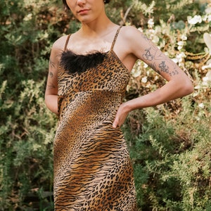 May include: A woman wearing a brown and black animal print slip dress with a black feather trim. The dress has a fitted silhouette and a short length. The woman is standing in front of a green leafy background.