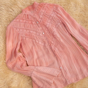 May include: A pink, long-sleeved blouse with a high collar and delicate lace detailing. The blouse has a button-up front and bell sleeves.