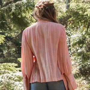 May include: A woman wearing a pink striped blouse with long sleeves and denim shorts. She is standing in a woodland setting with green trees and foliage.