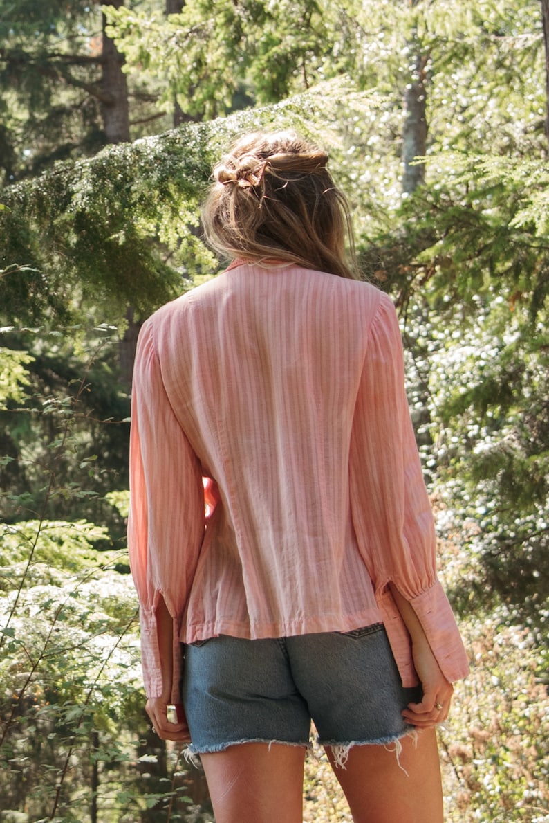 May include: A woman wearing a pink striped blouse with long sleeves and denim shorts. She is standing in a woodland setting with green trees and foliage.
