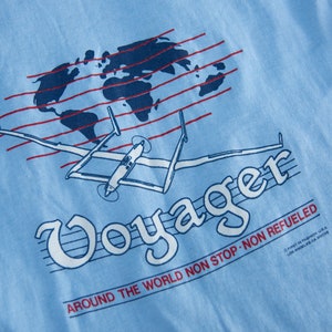 May include: Light blue t-shirt with a graphic of a world map, red lines, and a vintage airplane. The word "Voyager" is in a stylized font, with the text "AROUND THE WORLD NON STOP NON REFUELED".