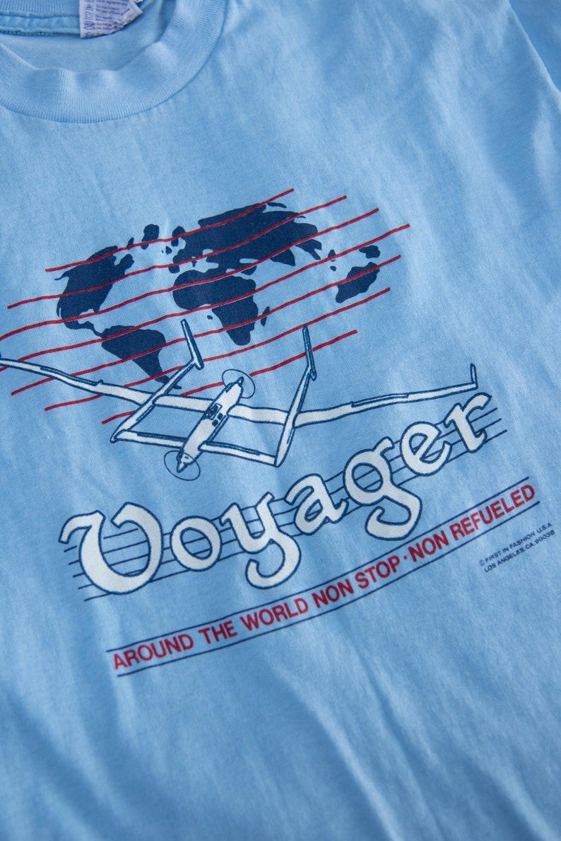 May include: Light blue t-shirt with a graphic of a world map, red lines, and a vintage airplane. The word "Voyager" is in a stylized font, with the text "AROUND THE WORLD NON STOP NON REFUELED".