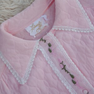 May include: A pink quilted robe with a white lace trim and floral embroidery. The robe has a collar and a button closure. The label on the robe reads "Leisure Life, Made in Hong Kong."