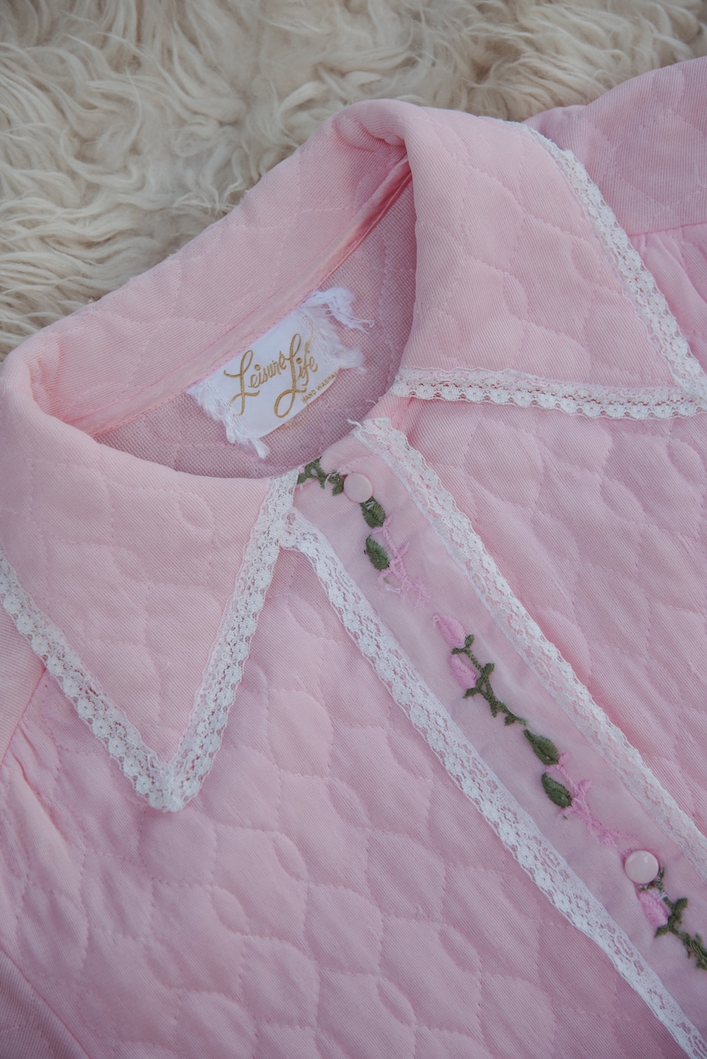 May include: A pink quilted robe with a white lace trim and floral embroidery. The robe has a collar and a button closure. The label on the robe reads "Leisure Life, Made in Hong Kong."
