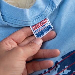 May include: Light blue t-shirt with a crew neck. The tag reads "NATIONAL FIFTY-FIFTY HEAVYWEIGHT" and "MADE IN/FAIT AU USA". The shirt has a graphic design with red and navy blue accents. The size is Large.