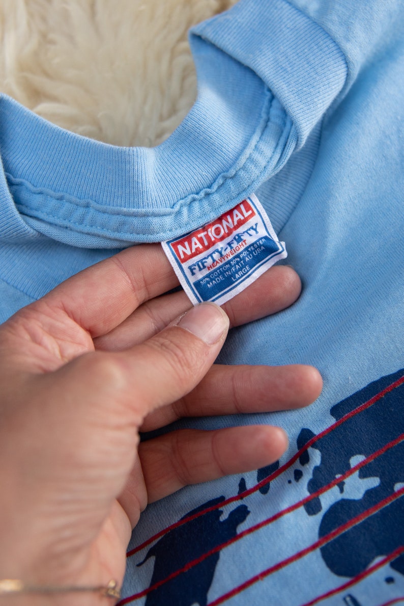 May include: Light blue t-shirt with a crew neck. The tag reads "NATIONAL FIFTY-FIFTY HEAVYWEIGHT" and "MADE IN/FAIT AU USA". The shirt has a graphic design with red and navy blue accents. The size is Large.