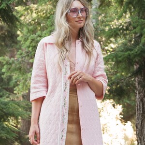 May include: A woman wearing a pink quilted jacket with floral embroidery and a brown skirt. She is wearing large, round, purple-tinted sunglasses.
