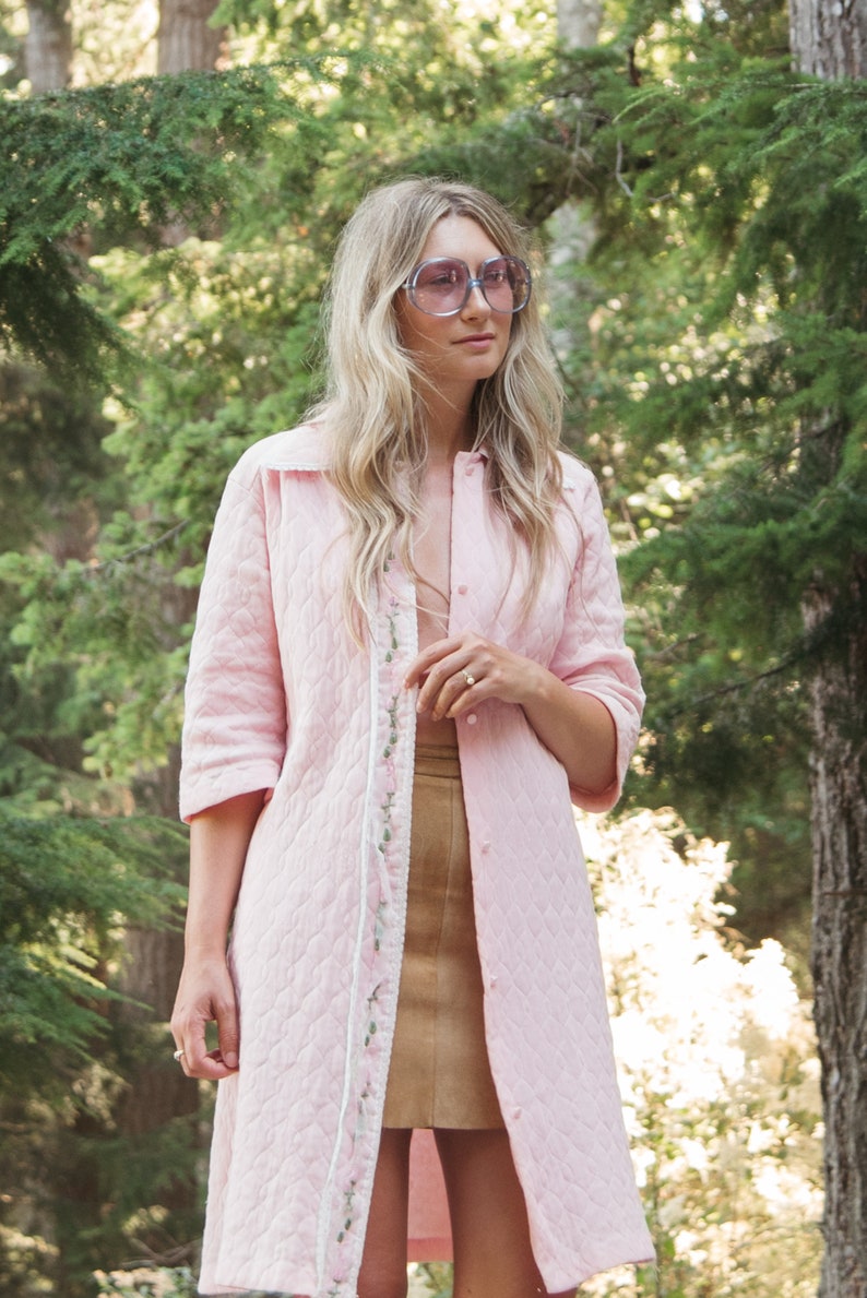 May include: A woman wearing a pink quilted jacket with floral embroidery and a brown skirt. She is wearing large, round, purple-tinted sunglasses.