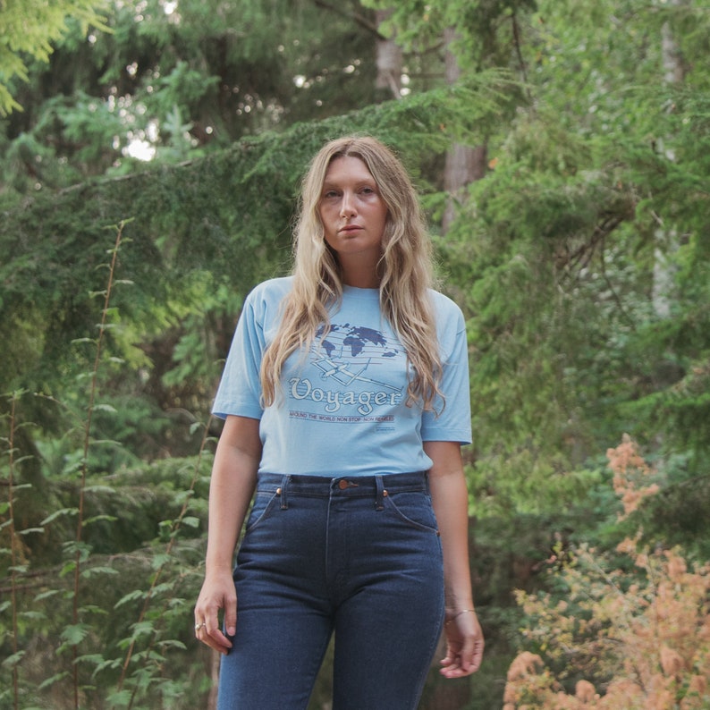 May include: A light blue t-shirt with the word "Voyager" and a world map graphic. The shirt is paired with dark blue jeans. The image is set against a backdrop of green trees.