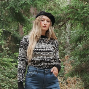 K&ouml;nnte beinhalten: Eine Frau tr&auml;gt einen schwarz-wei&szlig;en Pullover mit Muster, blaue Jeans und eine schwarze Baskenm&uuml;tze. Sie steht in einem Wald.