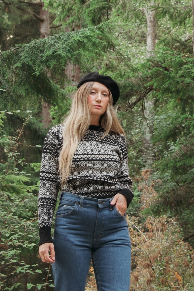 K&ouml;nnte beinhalten: Eine Frau tr&auml;gt einen schwarz-wei&szlig;en Pullover mit Muster, blaue Jeans und eine schwarze Baskenm&uuml;tze. Sie steht in einem Wald.