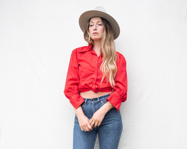 May include: A woman wearing a red button-down shirt tied at the waist, blue jeans, and a gray fedora.