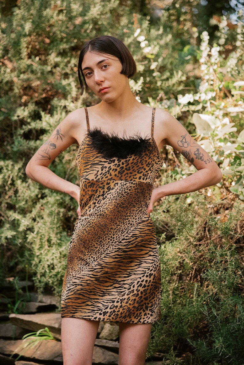 May include: A woman wearing a short, brown and black animal print dress with a black feather trim at the neckline. The dress has a fitted silhouette and a straight hemline. The woman is standing in front of a green leafy background.