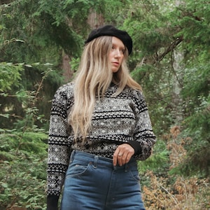K&ouml;nnte beinhalten: Eine Frau tr&auml;gt eine schwarze Baskenm&uuml;tze, einen wei&szlig;-schwarz gemusterten Pullover und eine blaue Jeans. Sie steht in einem Wald.