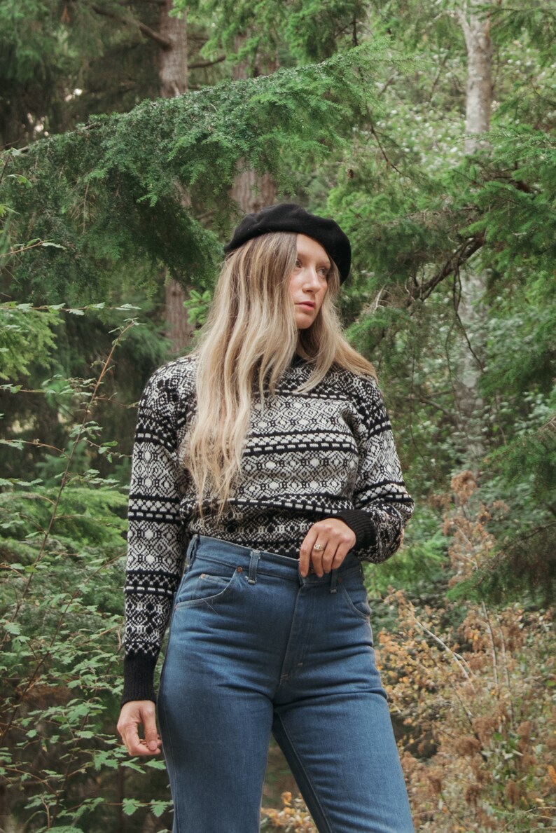 K&ouml;nnte beinhalten: Eine Frau tr&auml;gt eine schwarze Baskenm&uuml;tze, einen wei&szlig;-schwarz gemusterten Pullover und eine blaue Jeans. Sie steht in einem Wald.