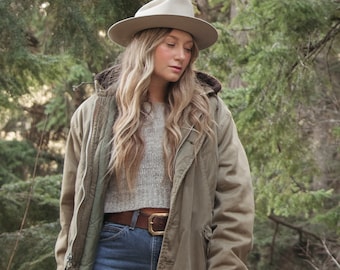 May include: A woman wearing a beige hat, a green jacket, a grey sweater, a brown belt, and blue jeans. She is standing in a forest.