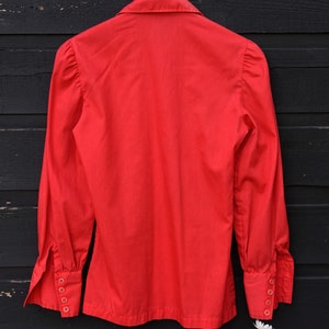 May include: A red long-sleeved button-up shirt with a collared neckline and puffed sleeves. The shirt has a classic design and is hanging on a wooden hanger against a black wooden background.