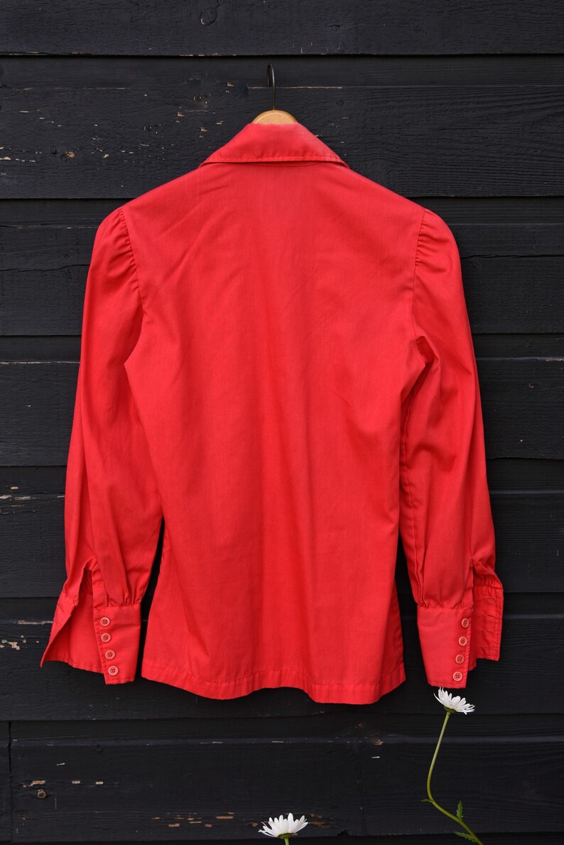 May include: A red long-sleeved button-up shirt with a collared neckline and puffed sleeves. The shirt has a classic design and is hanging on a wooden hanger against a black wooden background.