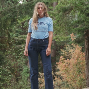 May include: A light blue t-shirt with the word "Voyager" and a world map graphic, paired with dark blue jeans and brown leather sandals. The outfit is set against a backdrop of lush green trees.