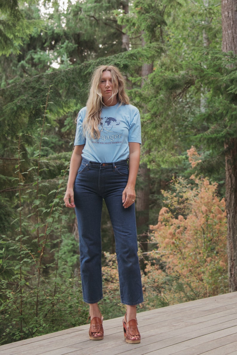 May include: A light blue t-shirt with the word "Voyager" and a world map graphic, paired with dark blue jeans and brown leather sandals. The outfit is set against a backdrop of lush green trees.