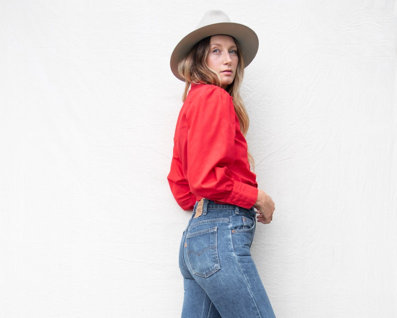 May include: A woman wearing a red collared shirt with long sleeves and blue jeans. She is wearing a white fedora hat.