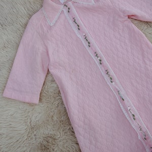 May include: A pink quilted robe with a white lace trim and green floral embroidery. The robe has a button-up front and short sleeves.