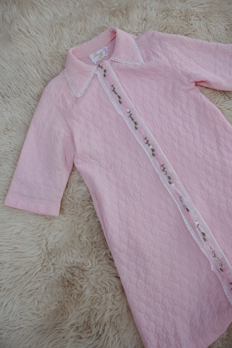 May include: A pink quilted robe with a white lace trim and green floral embroidery. The robe has a button-up front and short sleeves.