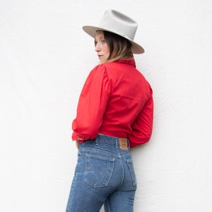 May include: A woman wearing a red button-down shirt with a white hat and blue jeans.