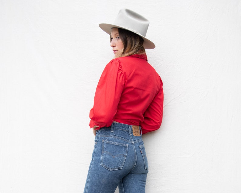 May include: A woman wearing a red button-down shirt with a white hat and blue jeans.