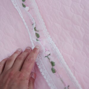 May include: A close-up of a pink fabric with a white lace trim and embroidered green leaves and pink flowers. The fabric has a quilted texture.