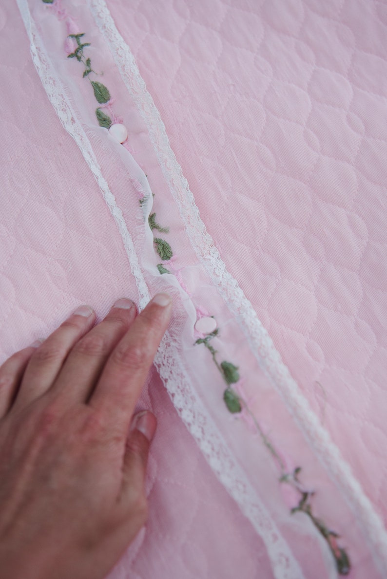 May include: A close-up of a pink fabric with a white lace trim and embroidered green leaves and pink flowers. The fabric has a quilted texture.