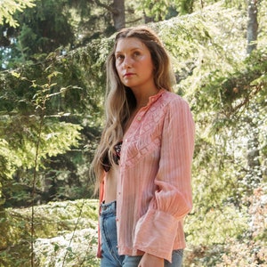 May include: A woman wearing a pink, embroidered, long-sleeved shirt with light blue denim shorts. She is standing in a woodland setting.