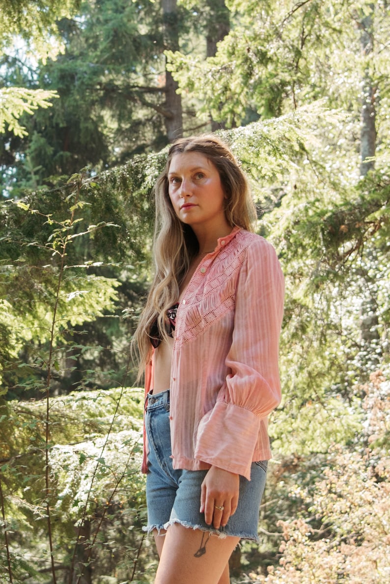 May include: A woman wearing a pink, embroidered, long-sleeved shirt with light blue denim shorts. She is standing in a woodland setting.