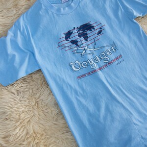 May include: Light blue t-shirt with a graphic of a world map, airplane, and the word "Voyager." The text "AROUND THE WORLD NON STOP - NON REFUELED" is also printed on the shirt. The shirt is made of a soft material.