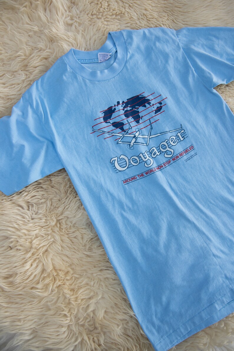May include: Light blue t-shirt with a graphic of a world map, airplane, and the word "Voyager." The text "AROUND THE WORLD NON STOP - NON REFUELED" is also printed on the shirt. The shirt is made of a soft material.