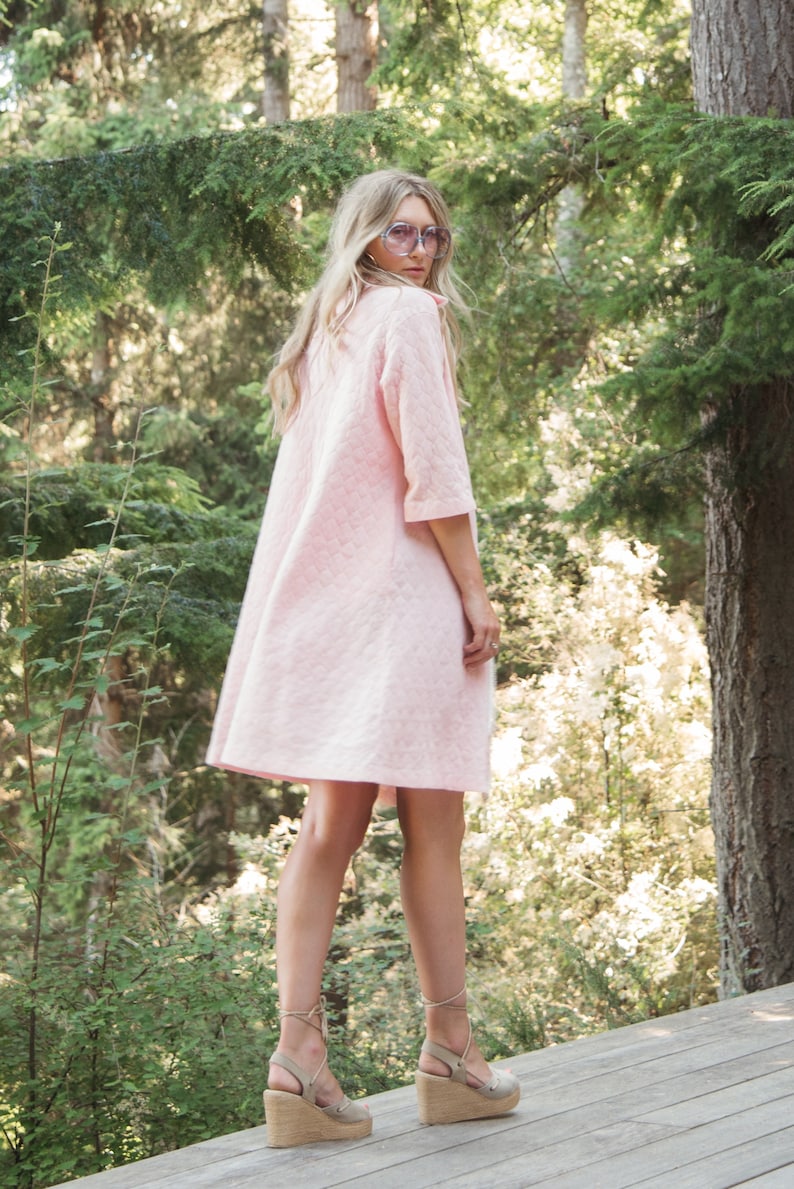 May include: A woman wearing a pink quilted dress and brown wedge sandals stands in a forest setting.