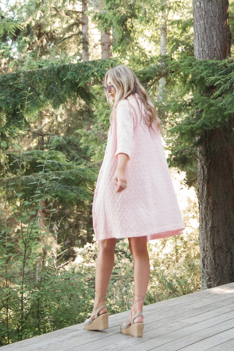 May include: A woman wearing a pink quilted dress and wedge sandals stands on a wooden deck in a forest setting.
