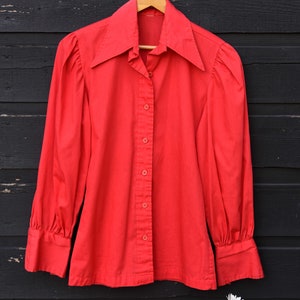 May include: A red long-sleeved button-up shirt with a large collar. The shirt is hanging on a wooden hanger against a black wooden background.