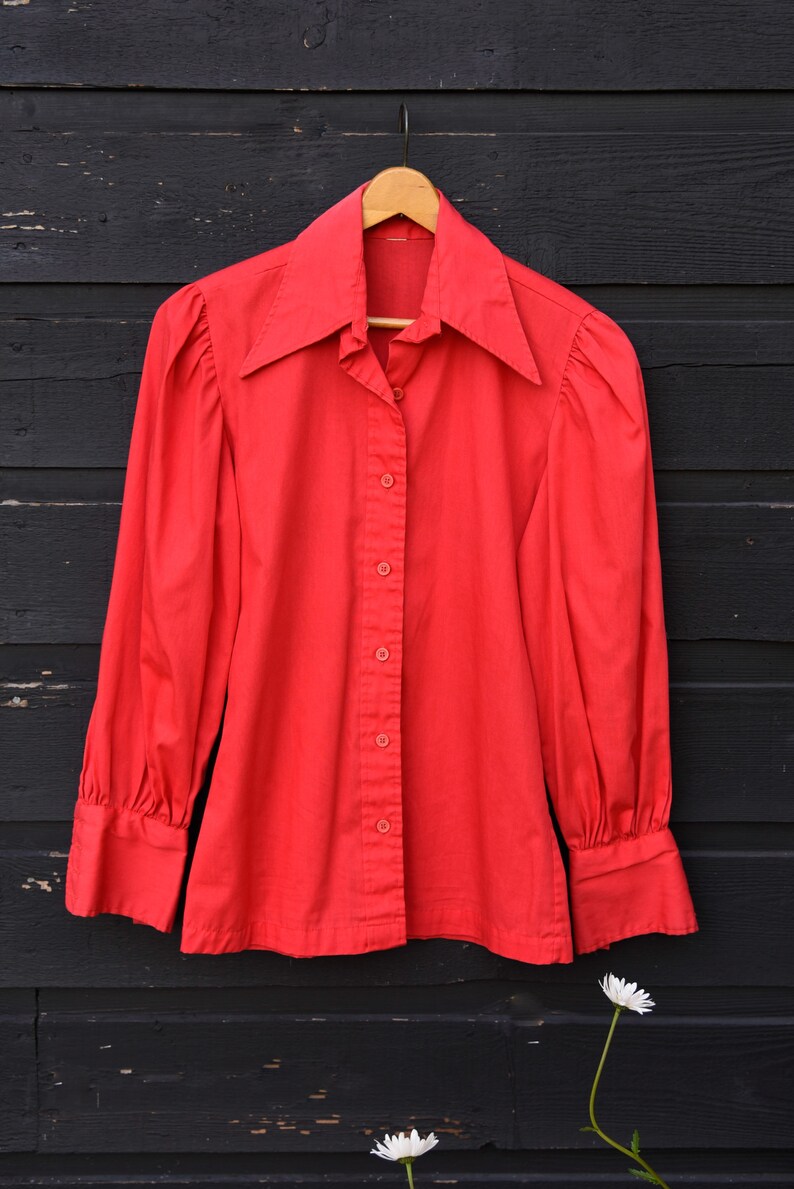 May include: A red long-sleeved button-up shirt with a large collar. The shirt is hanging on a wooden hanger against a black wooden background.