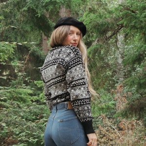 K&ouml;nnte beinhalten: Eine Frau tr&auml;gt einen schwarz-wei&szlig;en Pullover mit einem schwarzen Barett, blaue Jeans und einen Wald im Hintergrund.