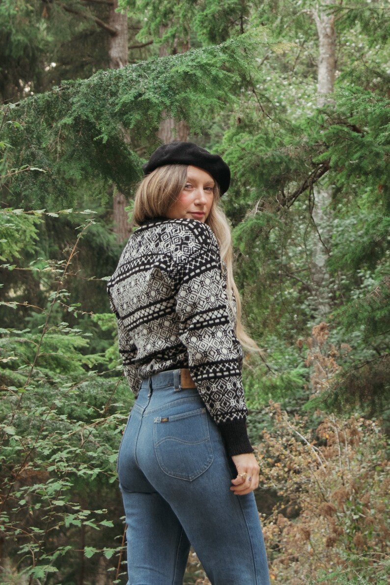 K&ouml;nnte beinhalten: Eine Frau tr&auml;gt einen schwarz-wei&szlig;en Pullover mit einem schwarzen Barett, blaue Jeans und einen Wald im Hintergrund.